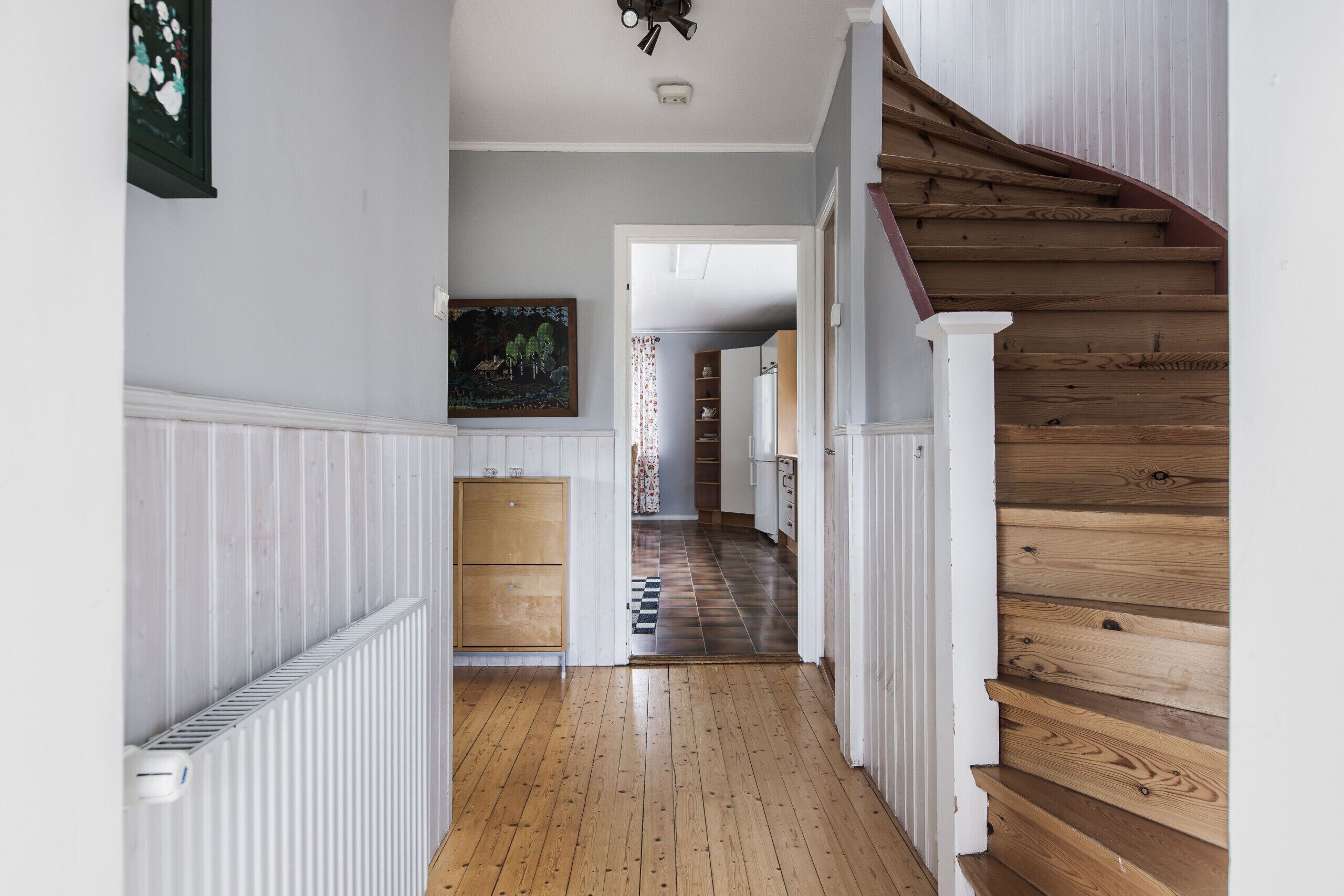Un pasillo con piso de madera, paredes blancas, un radiador, una escalera de madera y una puerta que da a otra habitación.