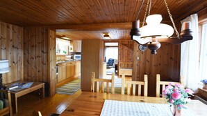 A wooden interior with a dining table, chairs, a vase with flowers, and a kitchen in the background.