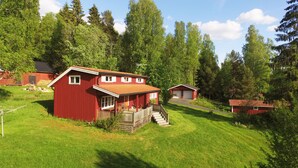 A red wooden house with a porch, surrounded by a grassy yard and tall trees.
