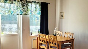 Een eethoek met een houten tafel en stoelen, een hanglamp, uitzicht op een meer door een raam, en een kleine ronde tafel met een metalen frame.