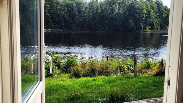 Uitzicht door een glazen deur op een houten terras met uitzicht op een meer, met weelderige begroeiing en bomen op de achtergrond.