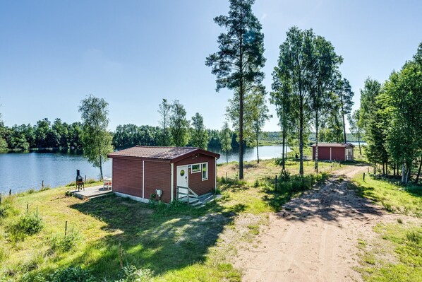 A small cabin with a red roof and white trim, situated near a lake with a dirt path leading to it. - Holiday home with lovely views of the lake Hängasjön | SE06015 (Hamneda)