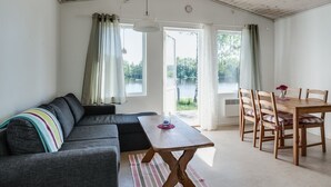 Ein gemütliches Wohnzimmer mit einer Couch, einem Holzesstisch und Stühlen. Große Fenster mit Vorhängen bieten Blick auf einen See und Bäume.