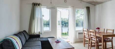 A cozy living room with a sofa, a wooden dining table, and chairs. There are large windows with curtains, and a view of a lake and trees outside.