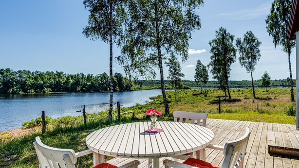 Ein runder Holztisch mit weißen Stühlen und roten Kissen, auf einer Holzterrasse mit Blick auf einen See.