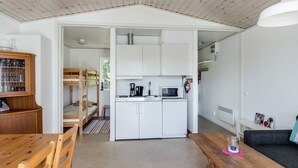 Eine kleine Küche mit weißen Schränken, einer Mikrowelle und einer Kaffeemaschine. Im Hintergrund steht ein Holztisch und ein Etagenbett.