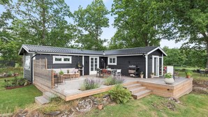 A black wooden cabin with a deck, a patio with outdoor furniture, and a garden area. - Fantastic cottage with panoramic views of Lake Vidöstern | SE06031 (Vittaryd)