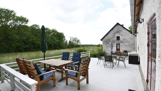 An outdoor patio with wooden furniture, a table, and chairs. There is a white building with a red door in the background. - Cozy and modern wing in Boge on Gotland's east coast | SE12011 (Slite)