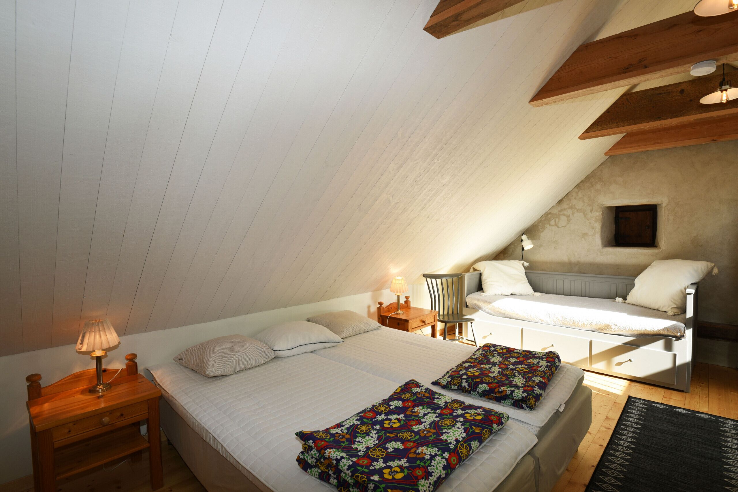 A bedroom with a sloped ceiling, a bed with floral bedding, a nightstand with a lamp, and a dresser.