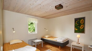Una habitación con dos camas, una mesita, un radiador, un ventanal con vistas a follaje y un cuadro de girasoles.