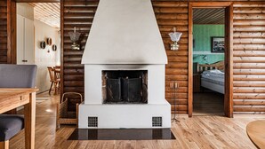 A log cabin interior with a central fireplace, wooden walls, a dining area, and a bedroom visible through an open door. - Cottage with its own sandy beach near VimmerbyI SE05018 (Vimmerby)