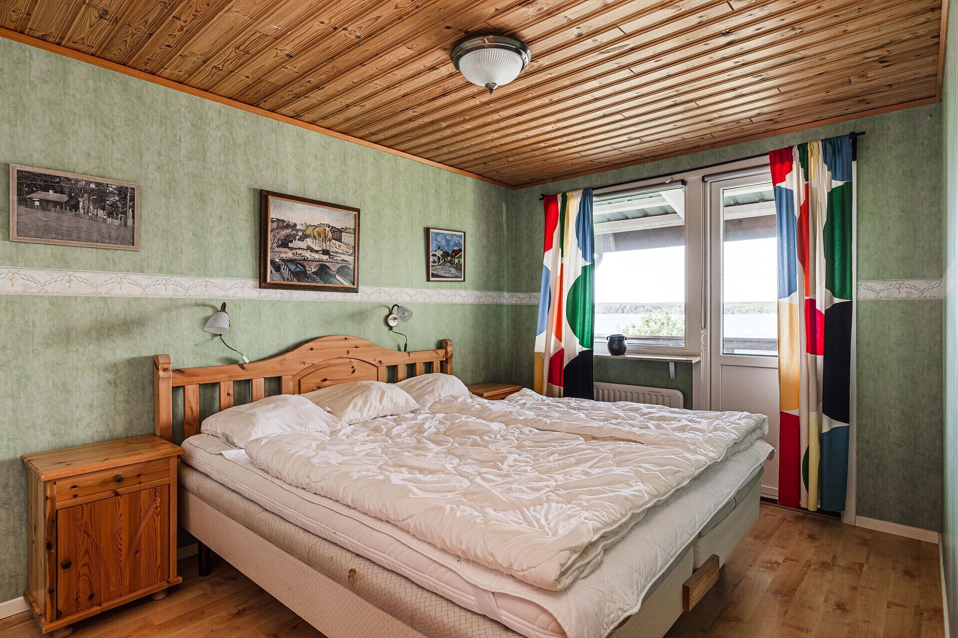 Een slaapkamer met een houten plafond, een bed met witte beddengoed, een houten nachtkastje en twee ingelijste foto’s aan de muur.