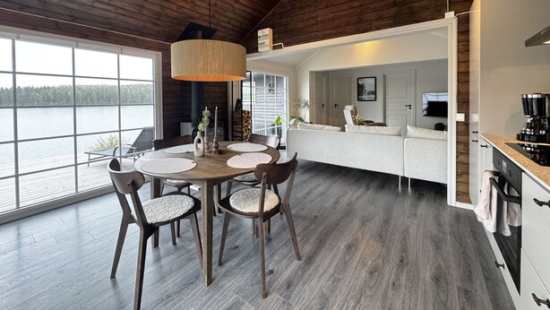 A modern interior with a dining area, kitchen, and living space, featuring wooden floors, a large window, and a view of a body of water.