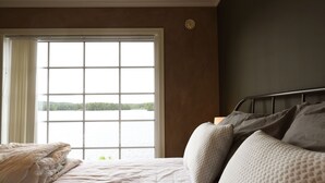 A bedroom with a large window, a bed with pillows, and a view of a lake.