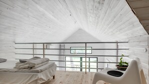 Une petite chambre mansardée, chaleureuse et accueillante, avec un plafond en pente, un lit dont la literie est soigneusement pliée, une chaise et une plante en pot.
