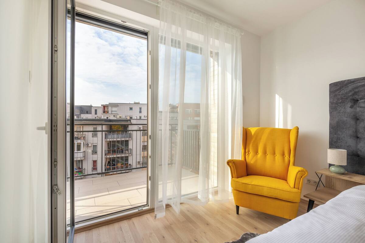 A room with a large sliding glass door, a yellow armchair, a bed, and a nightstand with a lamp.