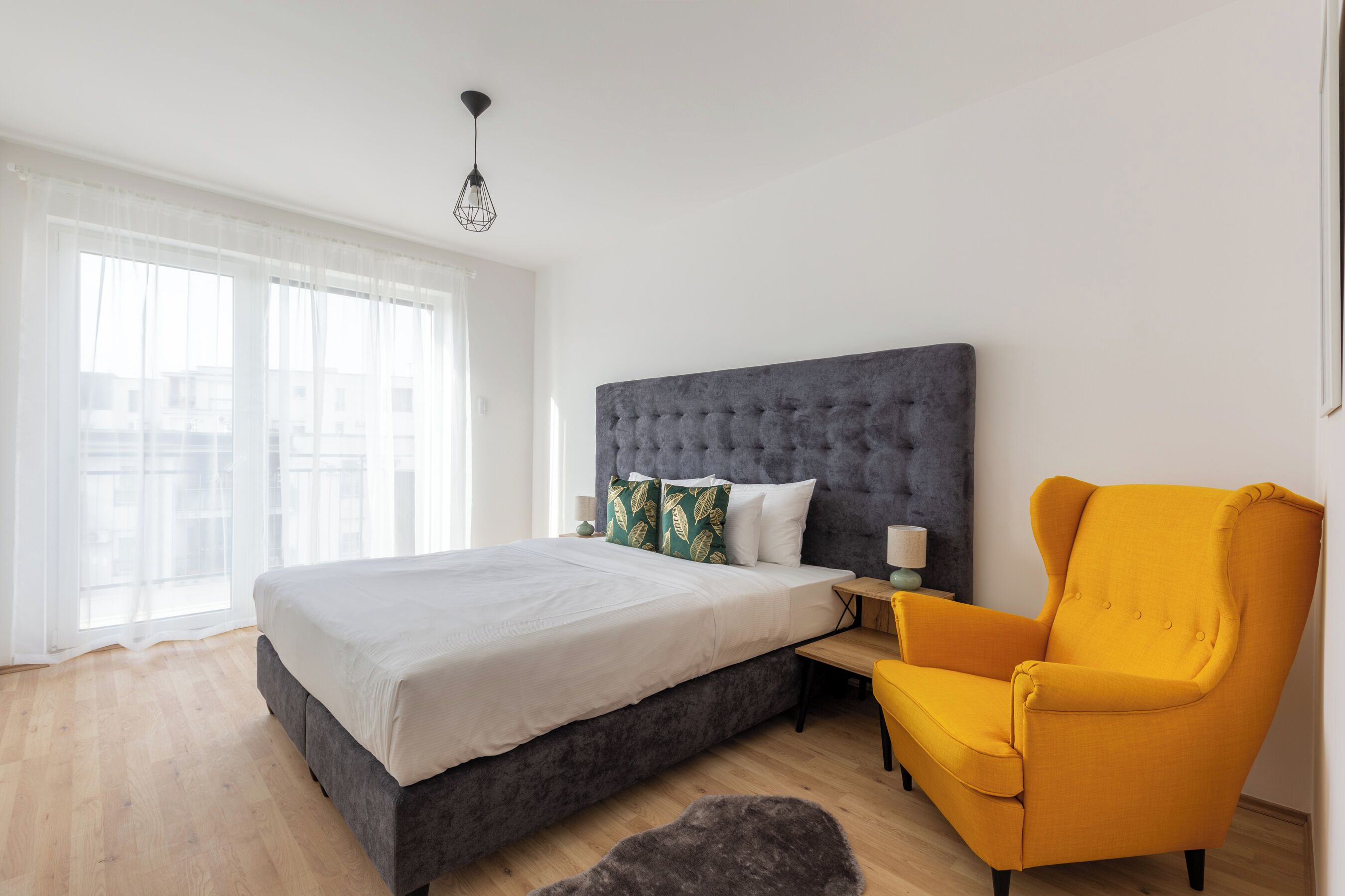 A modern bedroom with a large bed, a grey upholstered headboard, a yellow armchair, and a large window with sheer curtains.