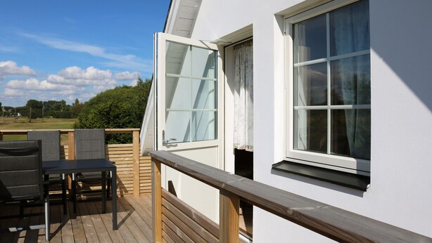 A white building with a wooden deck, a table with chairs, and a window with curtains. - Functional and cozy studio at Abbekås Golf Course | SE01024 (Abbekås)