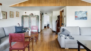A living room with a high ceiling, a wooden ceiling, a sofa, a red chair, a table, and a painting on the wall. - Nice holiday home near Skummelövs strand | SE02049 (Skummeslövsstrand)