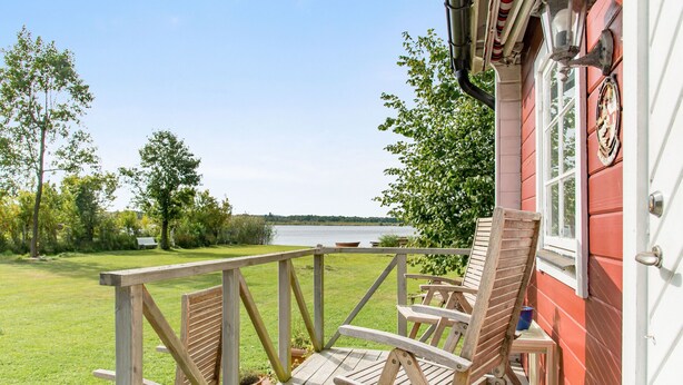 Eine Holzterrasse mit Bänken mit Blick auf einen See, ein rotes Gebäude mit einem weißen Fenster und ein klarer Himmel.