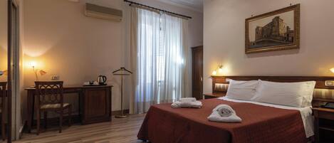 A hotel room with a bed, a desk, a chair, a lamp, a painting on the wall, and a window with curtains.