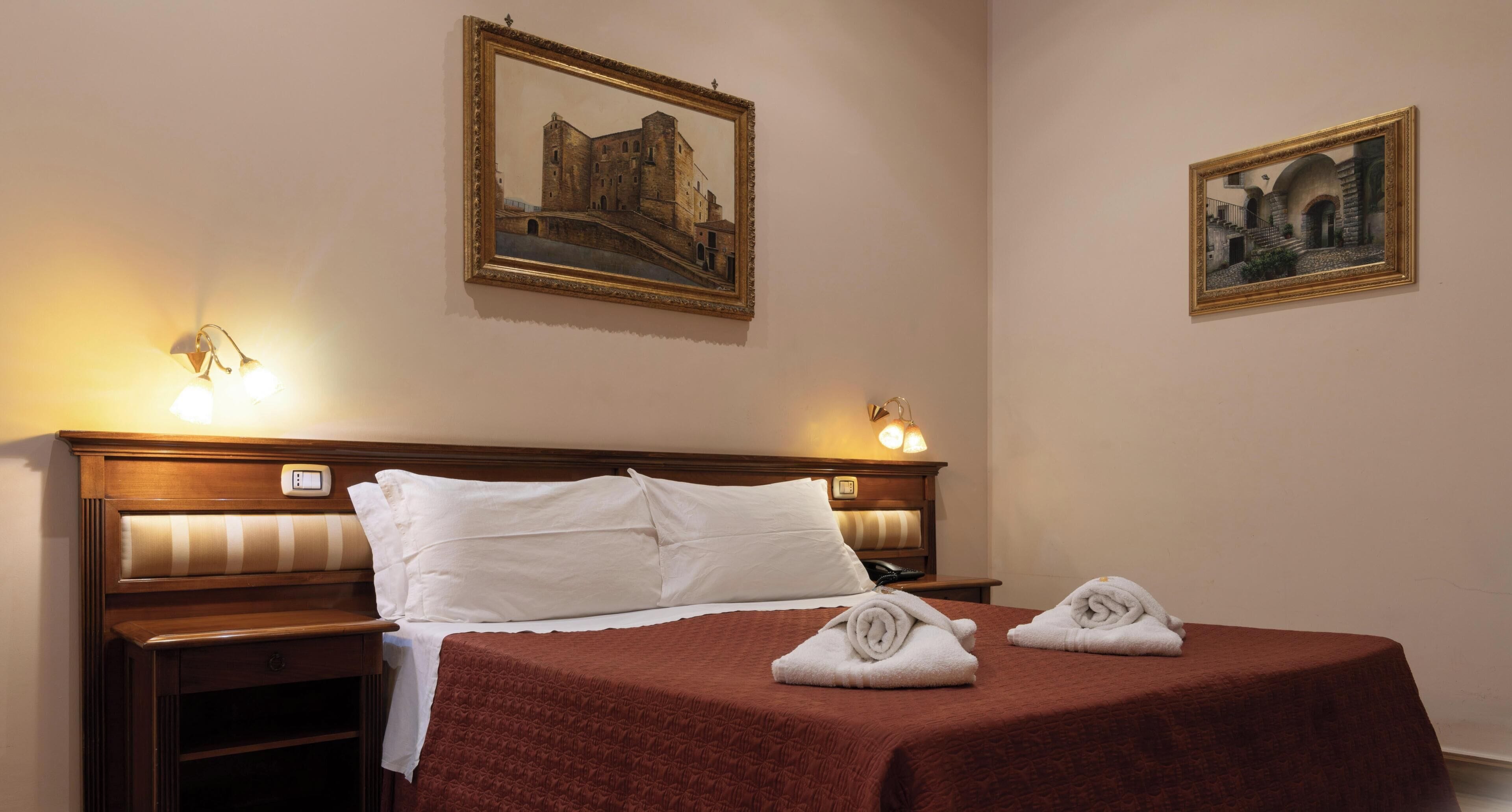 A hotel room with a bed, two bedside lamps, a nightstand, and two paintings on the wall.