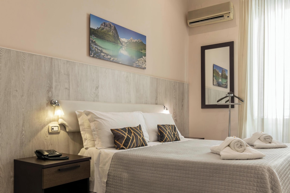 A hotel room with a bed, bedside table, lamp, and a painting of a mountain landscape.