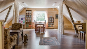 A spacious attic room with wooden slats on the ceiling, a dining table with chairs, two bookshelves, and a patterned rug on the floor. - Rural holiday home in Sjöared | SE02024 (Markaryd)