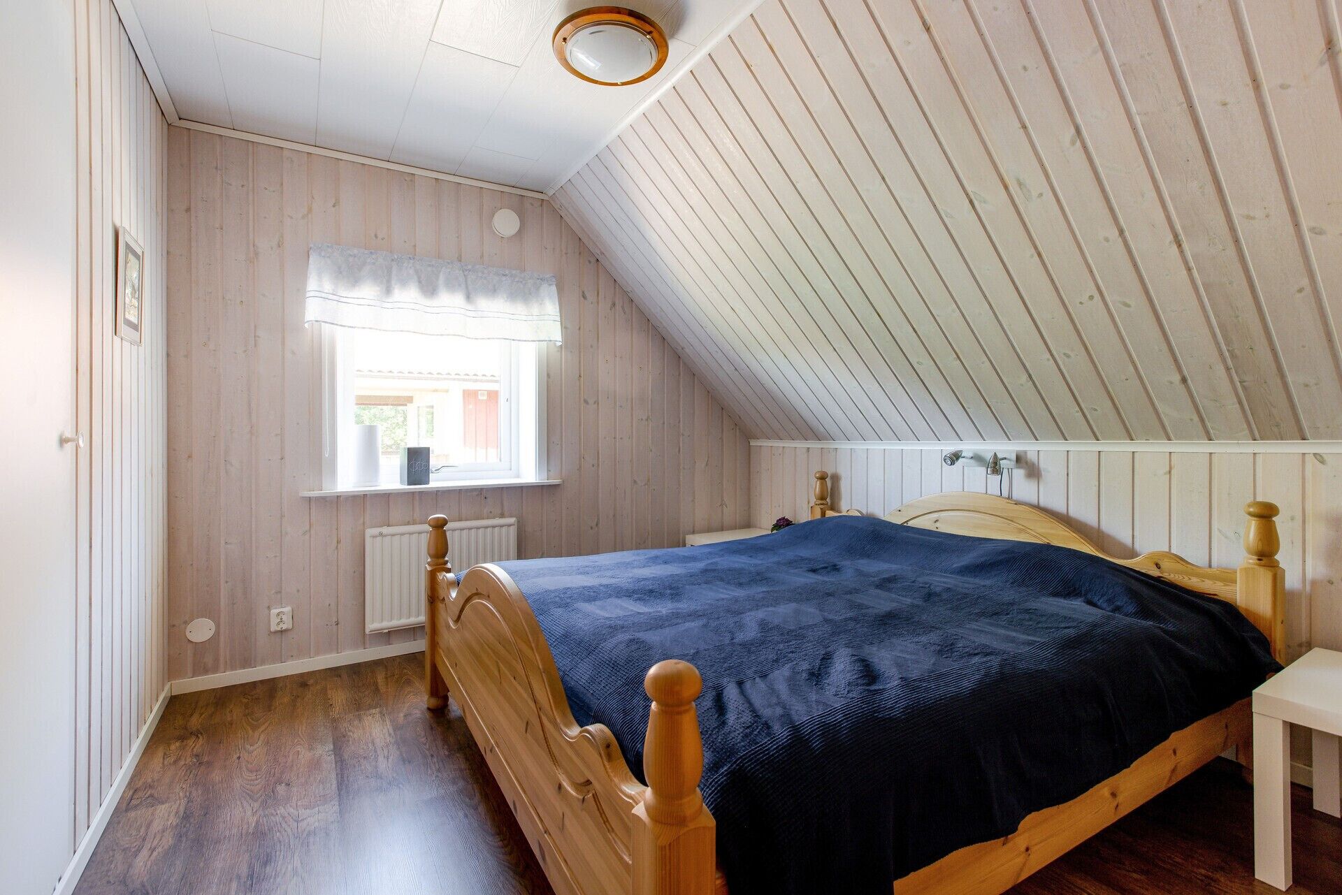 Ein Schlafzimmer mit schrägverdeckter Decke, Holzwänden, einem Bett mit blauer Bettdecke, einem Fenster mit weißem Vorhang und einem Heizkörper.
