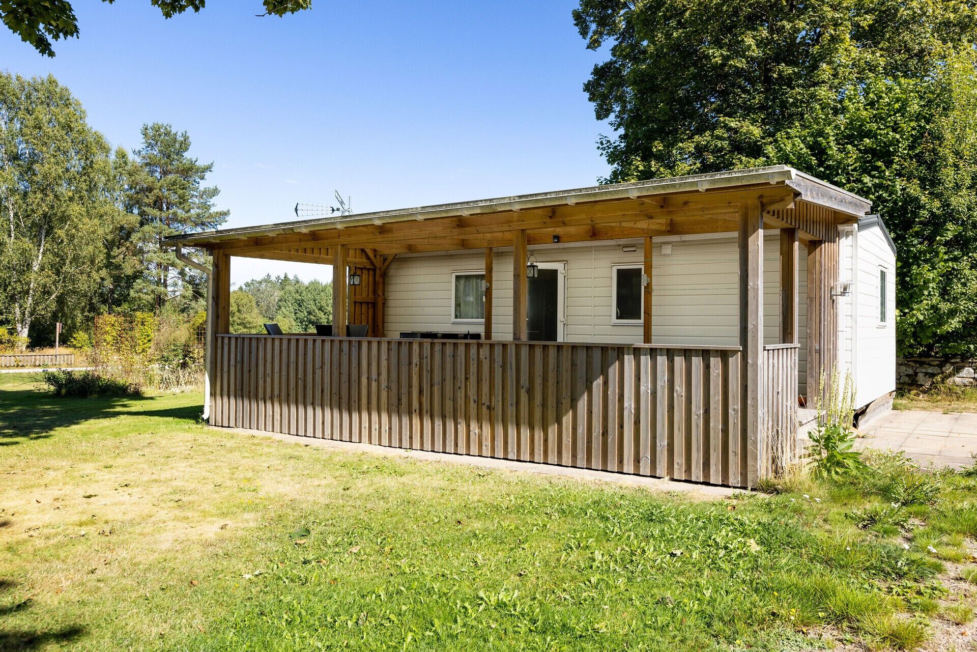 A small, single-story wooden cabin with a covered porch, a fenced yard, and a natural backdrop of trees and grass.