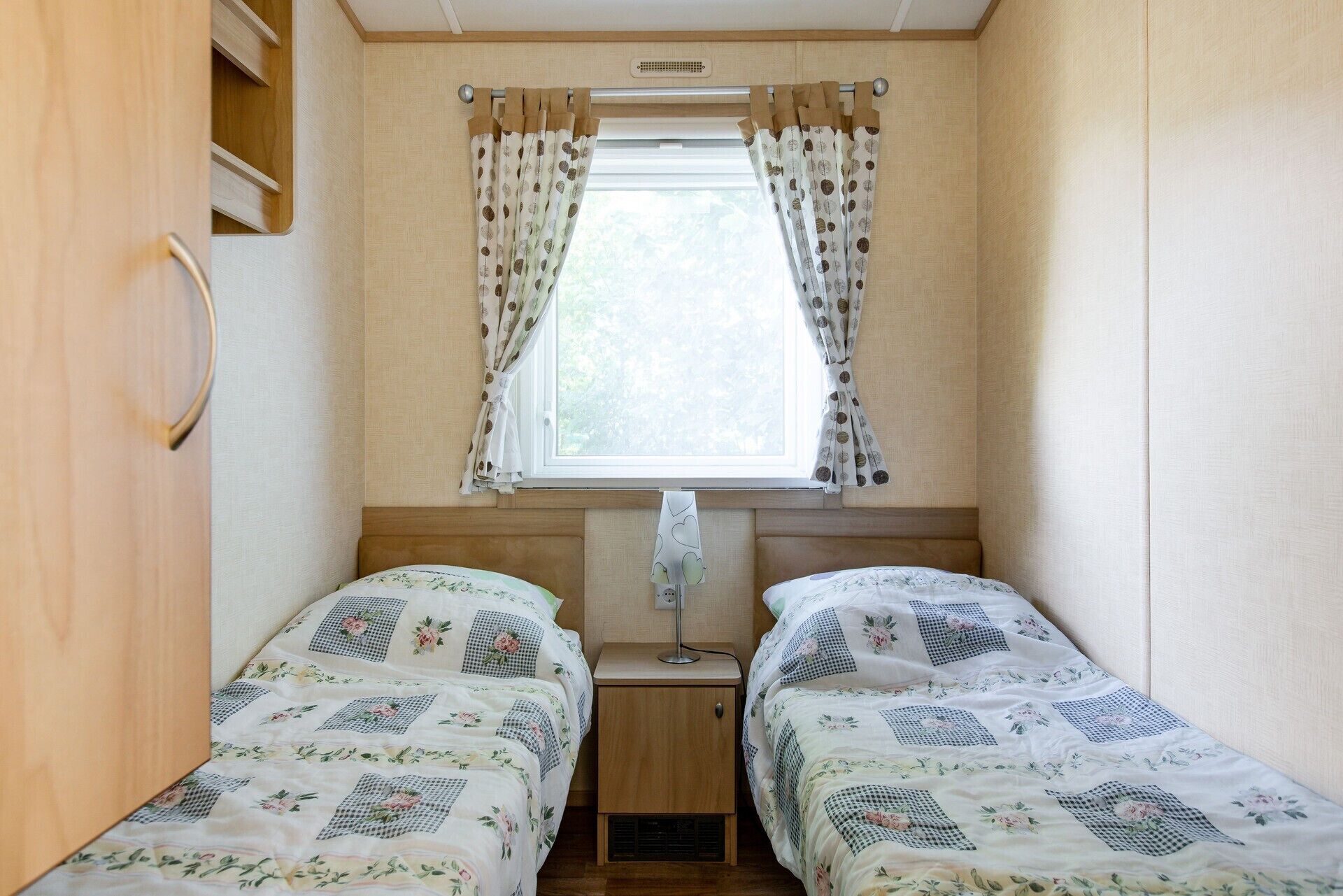 Une chambre compacte dans un camping-car, avec deux lits, une fenêtre avec des rideaux et une table de chevet avec une lampe.