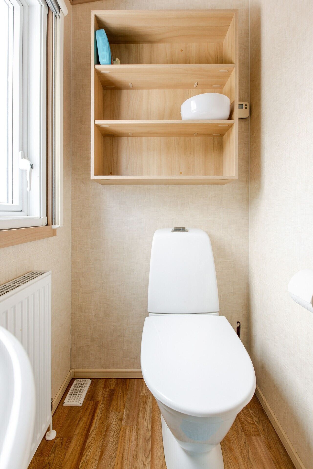 A bathroom with a white toilet, wooden shelves above it, and a window with blinds.