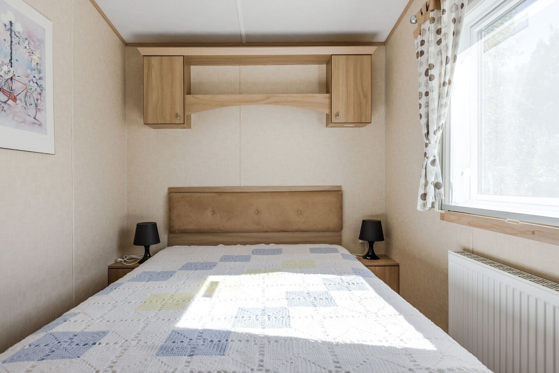 A bedroom with a wooden headboard, a quilted bedspread, two bedside tables with lamps, and a window with curtains.