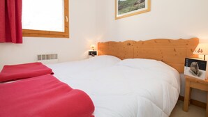 Une chambre à coucher avec une tête de lit en bois, une fenêtre ornée d’un rideau rouge, une table de chevet avec une lampe et un tableau encadré accroché au mur.