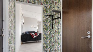 Un pasillo con papel tapiz estampado, una puerta de madera y una vista parcial de una habitación con cama y lámpara colgante.