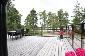 Una terraza de madera con una mesa y sillas, rodeada de barandillas de vidrio y plantas en macetas.
