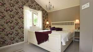 A bedroom with a white bed, floral wallpaper, a chandelier, and a window with curtains.