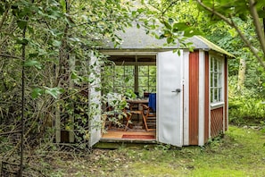 Un petit abri en bois, avec la porte ouverte, laissant apercevoir une table et des chaises à l’intérieur.
