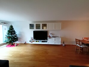 Un salon avec un téléviseur à écran plat, des meubles blancs, un plancher en bois et un sapin de Noël.