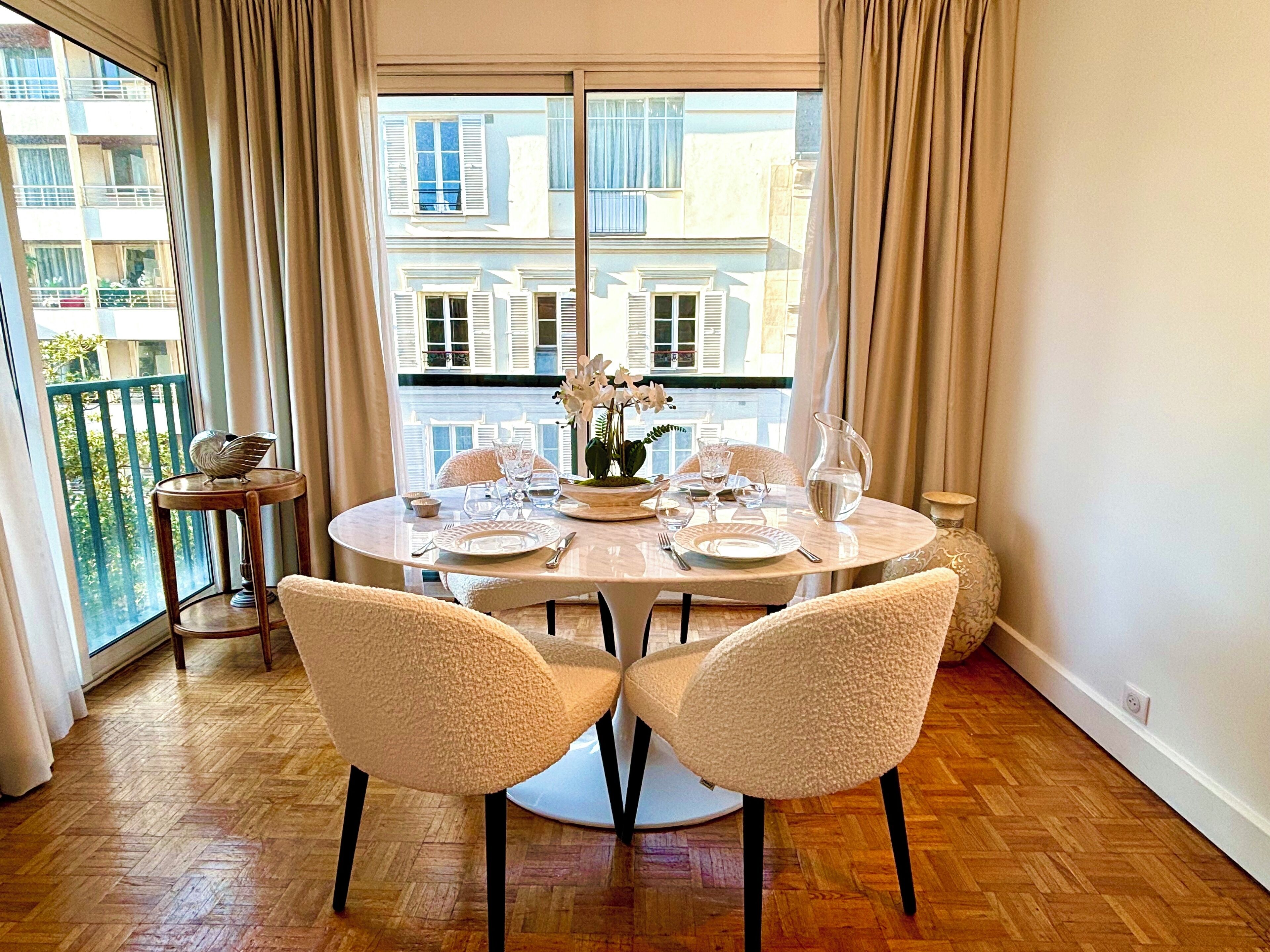 A dining area with a round table set for two, two chairs, a vase of flowers, and a view of buildings through large windows.