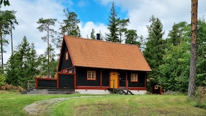 Punatiilikattoinen, mustaksi maalattu mökki, jossa on keltainen ovi. Sen ympärillä on puita, ja sen edessä on ruohikkoalue.