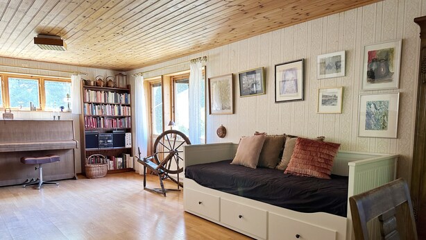 A cozy room with a wooden ceiling, a piano, a spinning wheel, a bed with storage drawers, a bookshelf, and framed pictures on the wall.