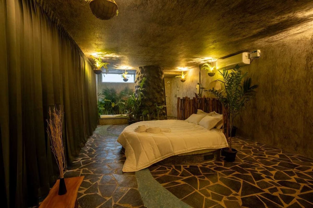 Una habitación con cama, suelo de piedra, plantas y una ventana.