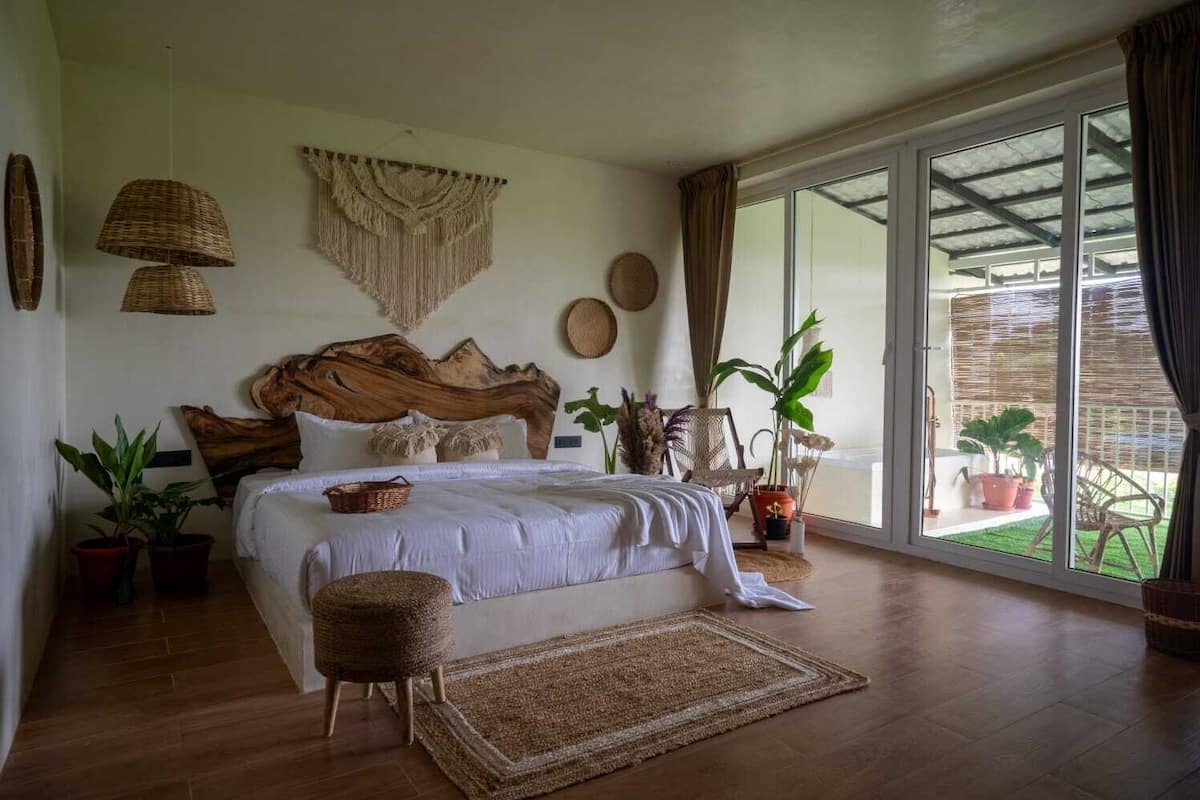 Un dormitorio espacioso con una cama grande, cabecero de madera y vistas a un jardín a través de amplias ventanas.