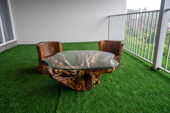 A glass-top table with a unique wooden base, surrounded by two wooden chairs, set on artificial grass.