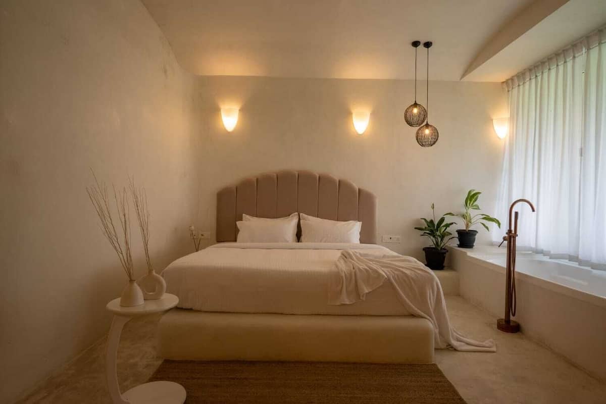 Un dormitorio con una cama grande, mesitas de noche, una planta en maceta y una ventana con cortinas.