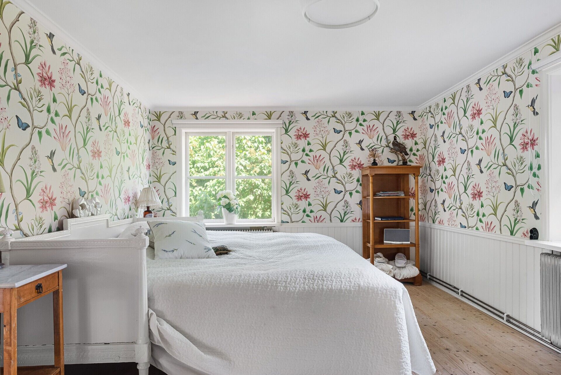 A bedroom with floral wallpaper, a bed, a bedside table, a lamp, a radiator, and a window with a view of greenery.