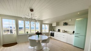 A modern kitchen with white cabinetry, a central island with chairs, a view of the sea through large windows, and a light blue refrigerator.