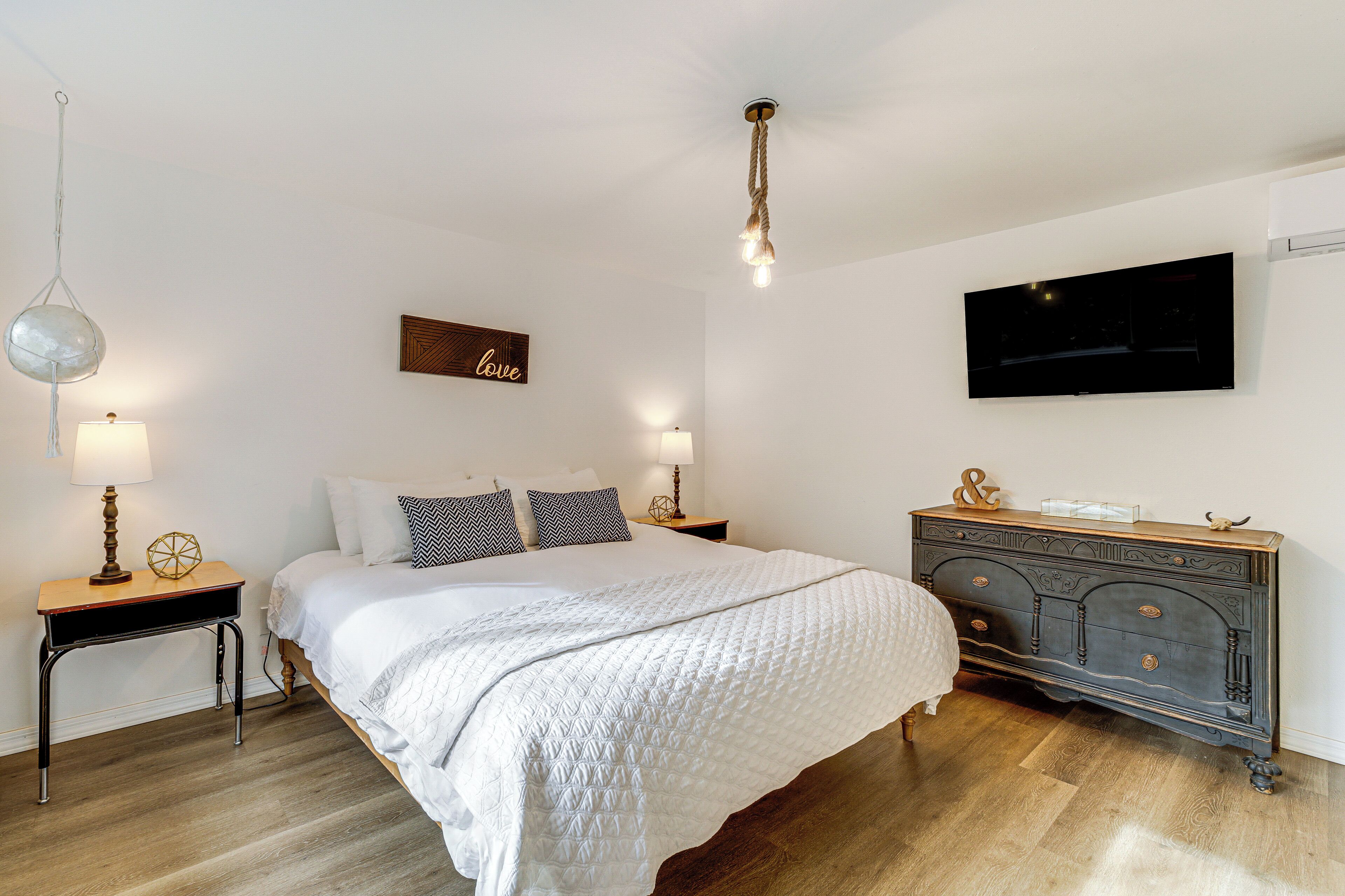 A bedroom with a large bed, two bedside tables with lamps, a dresser, a TV mounted on the wall, and a ceiling light fixture.