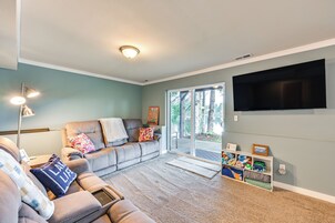 Una sala de estar con un sofá gris, una mesa de centro, un televisor de pantalla plana montado en la pared y un ventanal corredero que da a un espacio exterior.